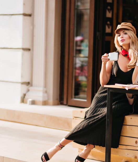 Attractive young woman resting after work in favorite cafe and enjoying coffee flavour. Outdoor portrait of blonde girl in stylish outfit relaxing in weekend.